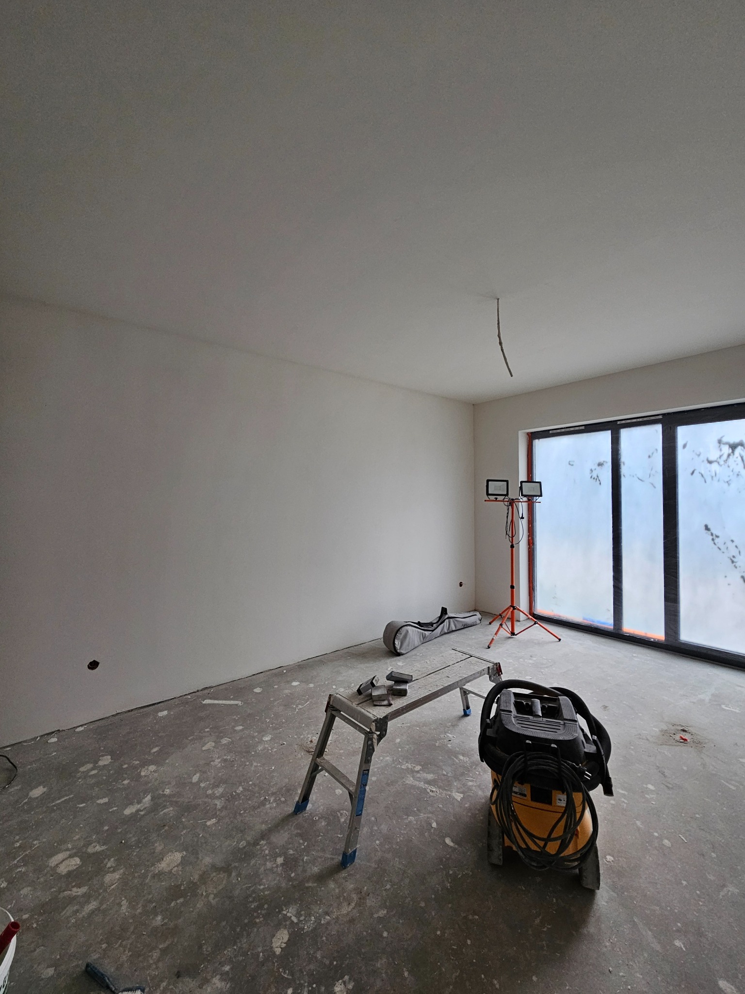 Pokój w trakcie prac wykończeniowych: świeżo pomalowane ściany, zakryte okna, drabina, lampy budowlane i odkurzacz przemysłowy na zalanym farbą betonie.