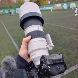 BABCZYŃSKI IT Piotr Babczyński - Ręka trzyma aparat Sony z teleobiektywem 100-400 mm G OSS, mikrofon na sankach, w tle boisko piłkarskie z grającymi zawodnikami