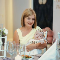 BABCZYŃSKI IT Piotr Babczyński - Kobieta w żółtej sukience trzyma na rękach niemowlę w białej, koronkowej sukience z kwiatowymi aplikacjami podczas uroczystości, widoczne dekoracje balonowe i zastawa stołowa.