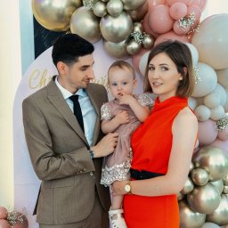 BABCZYŃSKI IT Piotr Babczyński - Uśmiechnięta rodzina z małą dziewczynką w sukience na tle dekoracji z balonów w odcieniach różu i złota.