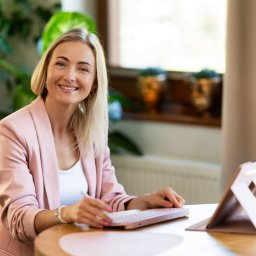 BABCZYŃSKI IT Piotr Babczyński - Uśmiechnięta blond kobieta w różowym żakiecie pracuje na tablecie z klawiaturą, używając rysika. W tle jasne okno i zielona roślina.