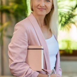 BABCZYŃSKI IT Piotr Babczyński - Portret uśmiechniętej blondynki w różowej marynarce, trzymającej jasnoróżowy notatnik z zakładką, na tle zieleni.