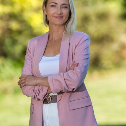 BABCZYŃSKI IT Piotr Babczyński - Portret uśmiechniętej kobiety w różowym żakiecie i białych spodniach, z rękami skrzyżowanymi na tle zieleni.