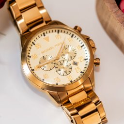 BABCZYŃSKI IT Piotr Babczyński - Złoty zegarek Michael Kors na białym tle, z drewnianym pojemnikiem wypełnionym różowymi różami w tle.