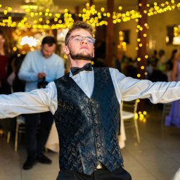 BABCZYŃSKI IT Piotr Babczyński - Mężczyzna w czarnej kamizelce i muszce, z rozłożonymi ramionami, na tle sali weselnej z dekoracjami świetlnymi i gośćmi.