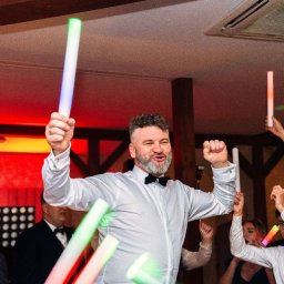 BABCZYŃSKI IT Piotr Babczyński - Uczestnicy wesela z podniesionymi świetlnymi pałeczkami, mężczyzna w białej koszuli i muszce w centrum kadru, w tle osoba filmująca kamerą.