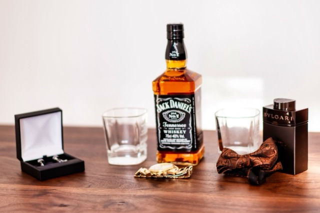 Przygotowania do ślubu: butelka Jack Daniel's, dwa szklane kieliszki, brązowa muszka, perfumy Bvlgari i spinki do mankietów w czarnym pudełku, ułożone na drewnianym stole.