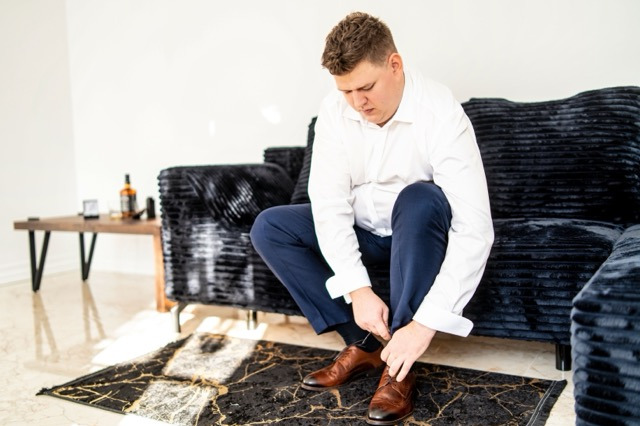 Mężczyzna w białej koszuli i granatowych spodniach, klęczący na podłodze z czarnym dywanem w złote żyłki, zawiązuje sznurowadła w brązowych skórzanych butach przed czarną, pluszową sofą. Na stoliku...