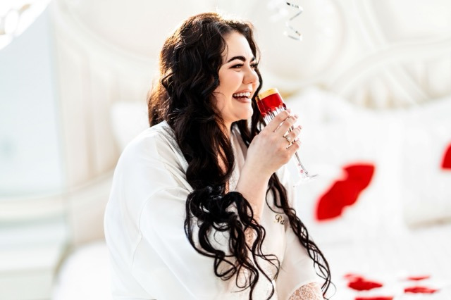 Uśmiechnięta panna młoda w białym szlafroku z koronkowymi rękawami, trzymająca kieliszek z czerwonym płynem, w tle białe łóżko z czerwonymi sercami i srebrne balony.