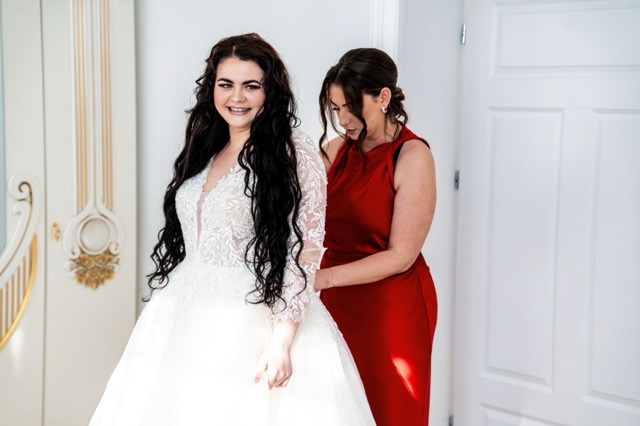 Uśmiechnięta panna młoda w koronkowej sukni z długimi rękawami, dopinana przez kobietę w czerwonej sukience na tle białych drzwi i dekoracyjnego panelu ściennego.