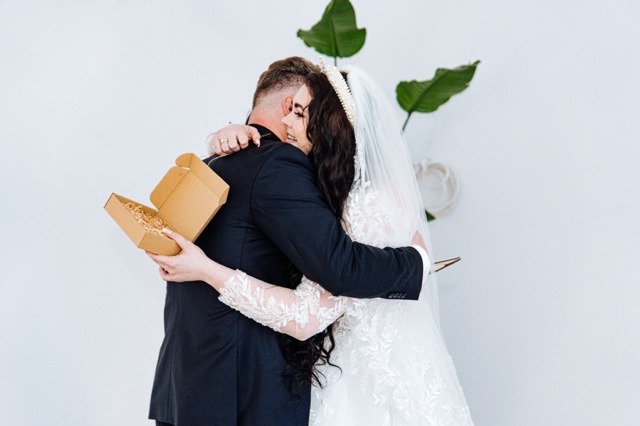 Para młoda w objęciach, panna młoda w koronkowej sukni i welonie, pan młody w ciemnym garniturze trzymający otwarte pudełko z siankiem dekoracyjnym.