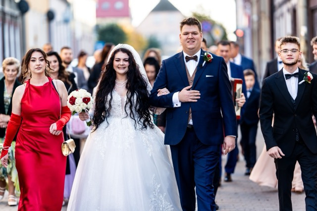 Para młoda w trakcie marszu z gośćmi weselnymi ulicą miejską; panna młoda w białej sukni z bukietem czerwono-białych róż, pan młody w granatowym garniturze z bordową muchą.