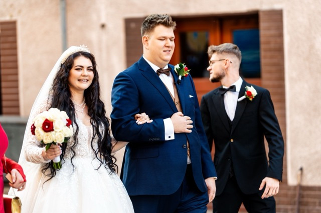 Uśmiechnięta panna młoda w koronie i z bukietem czerwono-białych róż, idąca pod rękę z panem młodym w granatowym garniturze i brązową muchą, obok mężczyzna w czarnym garniturze z muchą.