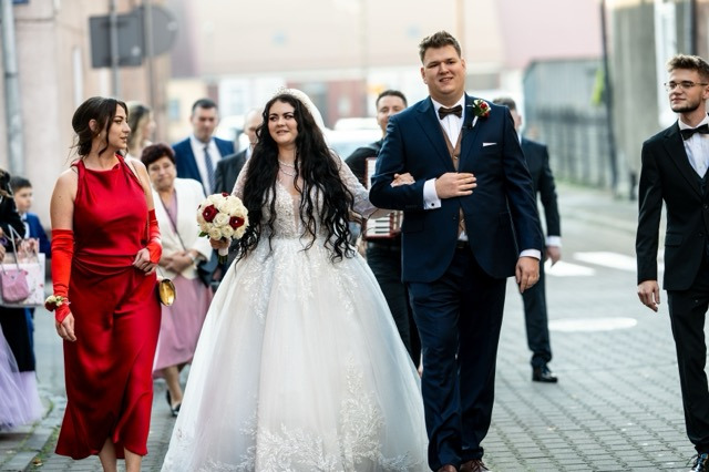 Para młoda idzie ulicą w otoczeniu gości weselnych; panna młoda w sukni z koronką, pan młody w granatowym garniturze z bordową muchą.