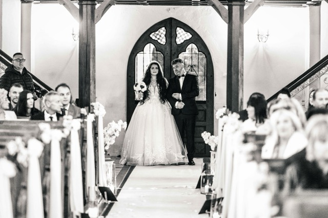 Czarno-białe zdjęcie panny młodej w sukni z koronkowymi zdobieniami prowadzonej do ołtarza przez mężczyznę w smokingu, na tle gotyckich drzwi i rzędów gości.