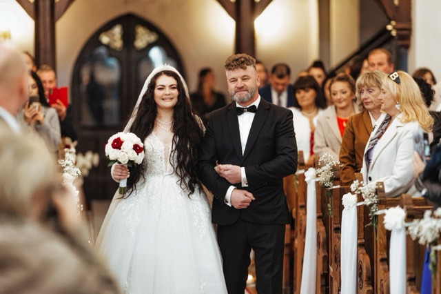 Panna młoda w białej sukni z bukietem czerwono-białych róż, prowadzona do ołtarza przez ojca w czarnym smokingu, w tle goście i zdobione ławki kościelne.