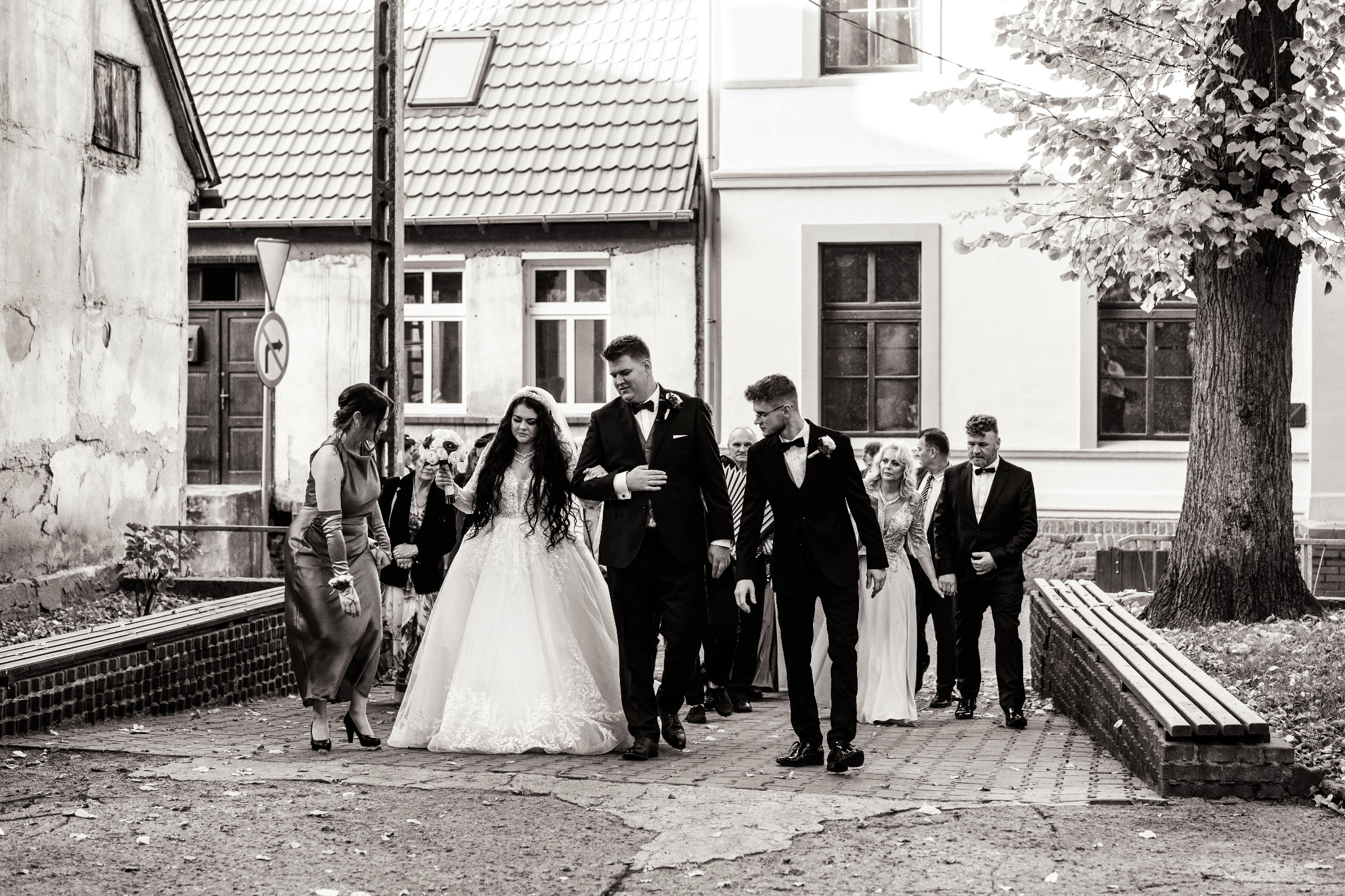 Czarno-białe zdjęcie: Para młoda w towarzystwie gości weselnych idzie brukowaną uliczką między budynkami.