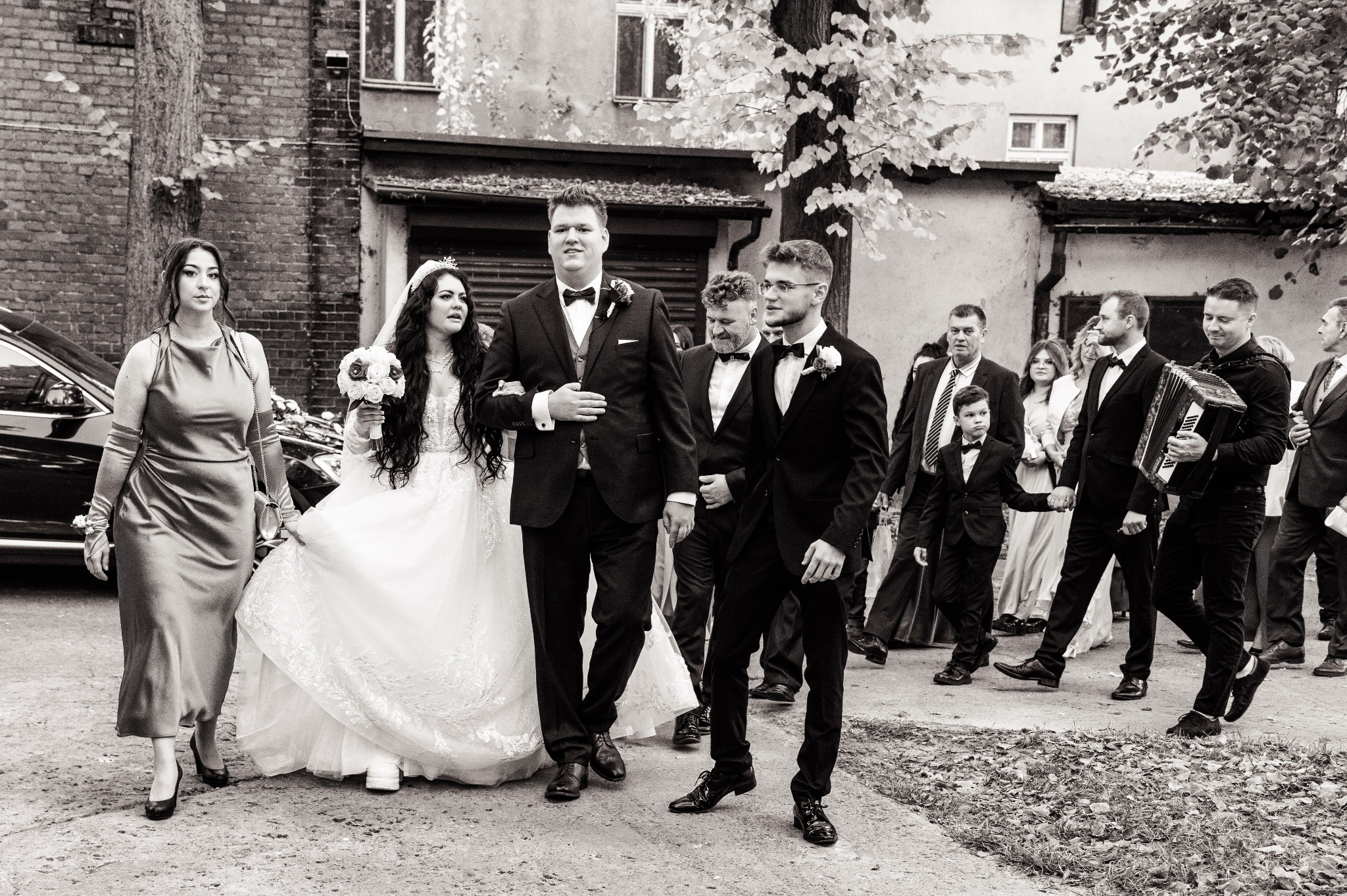 Czarno-białe zdjęcie grupy osób wychodzących z budynku, w tym panny młodej w koronie i sukni z koronkowymi zdobieniami, orszaku weselnego i akordeonisty.