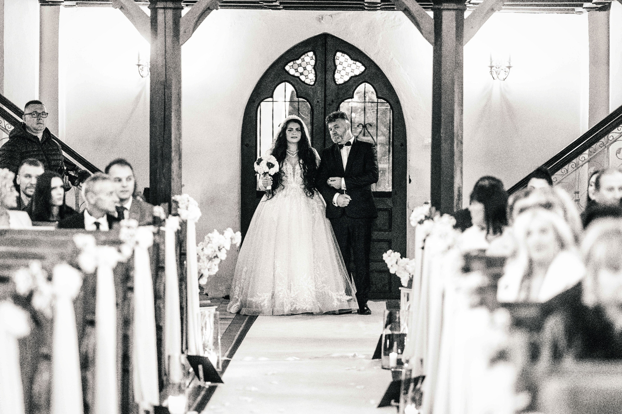 Czarno-białe zdjęcie panny młodej w koronkowej sukni i pana młodego w smokingu, prowadzonych przez ojca do ołtarza, w tle gotyckie drzwi i goście.