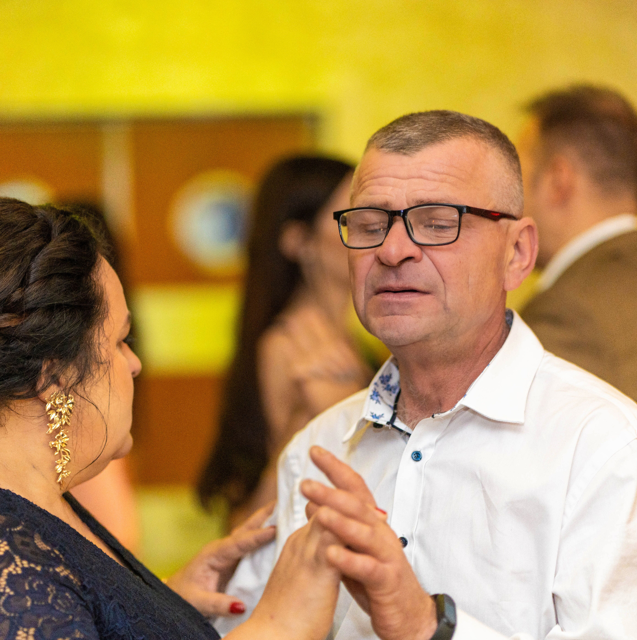 Para w średnim wieku, on w okularach i białej koszuli z niebieskimi guzikami, ona w granatowej koronkowej sukience i złotych kolczykach, tańczą blisko siebie na tle żółtej ściany.