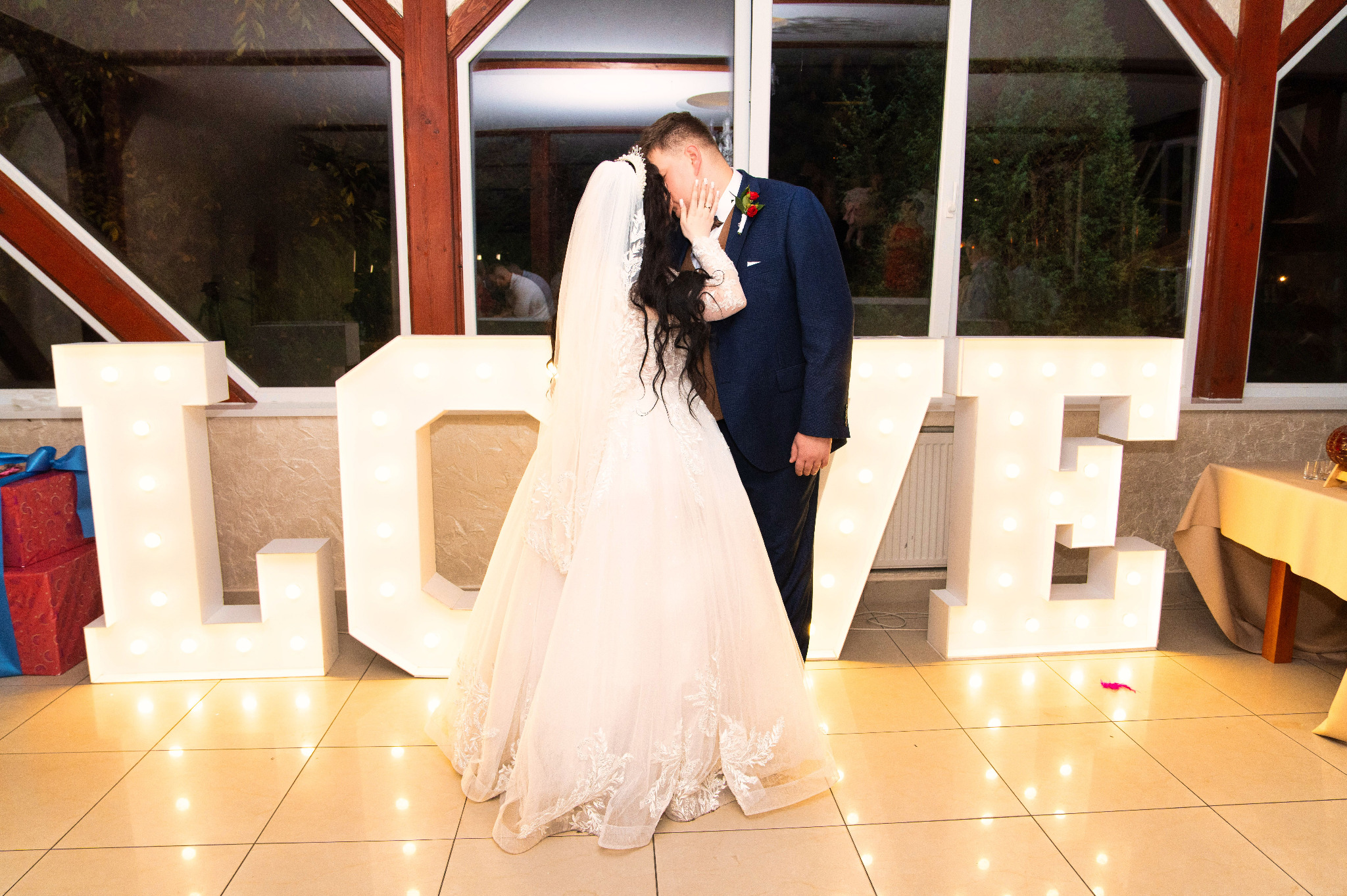 Para młoda w objęciach, panna młoda w białej sukni z koronkowymi zdobieniami i długim welonem, pan młody w granatowym garniturze, na tle dużych, podświetlanych liter LOVE