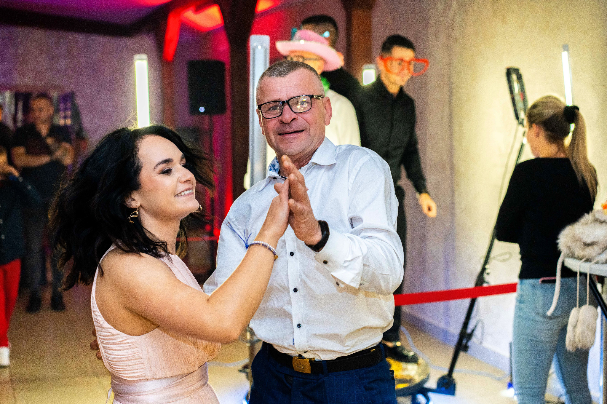 Uśmiechnięta para tańczy na parkiecie podczas eventu, w tle osoby z rekwizytami i oświetlenie.