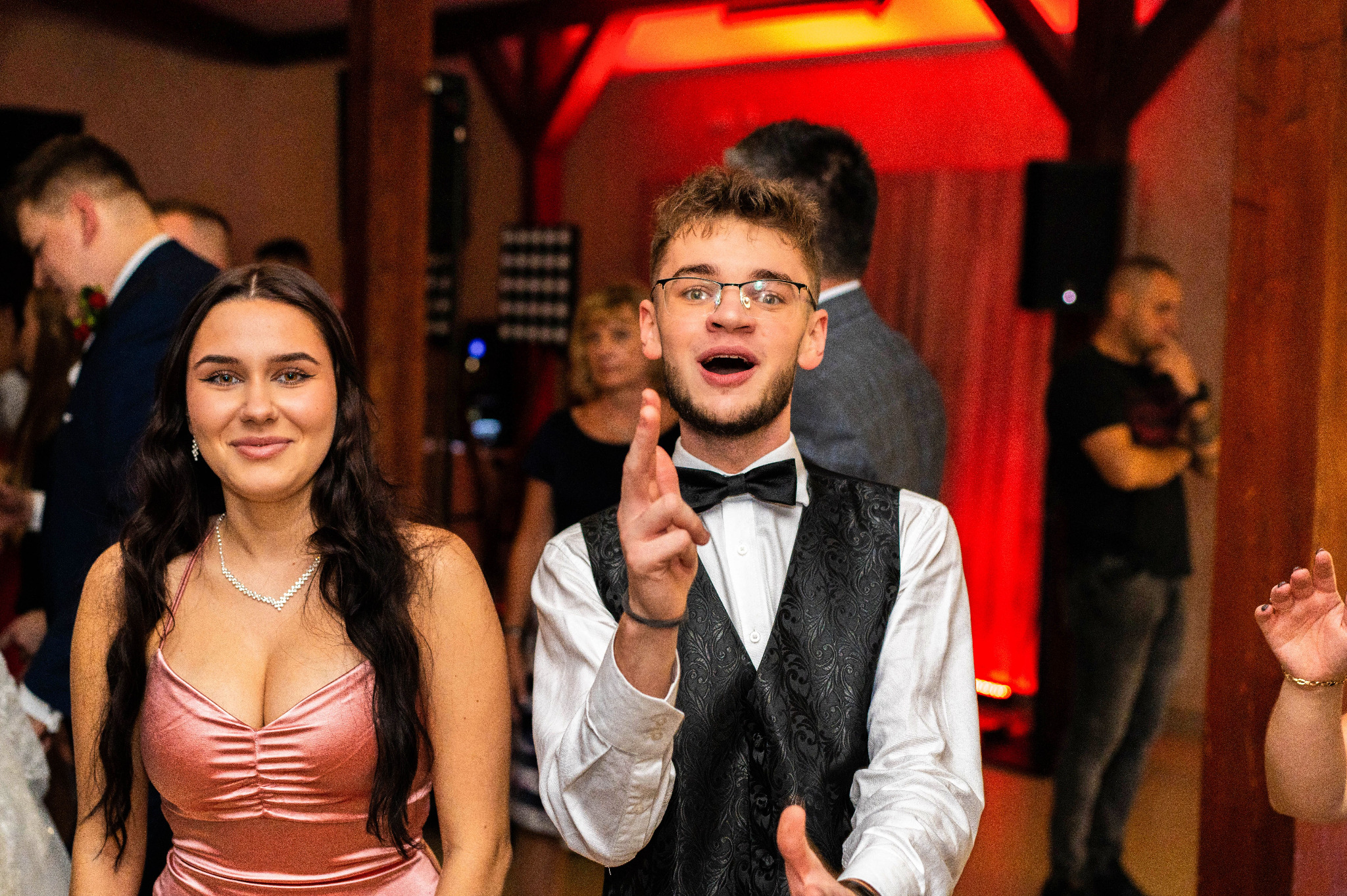 Uśmiechnięta kobieta w różowej sukience i mężczyzna w muszce i kamizelce gestykulujący na parkiecie podczas wydarzenia, w tle goście i czerwone oświetlenie.