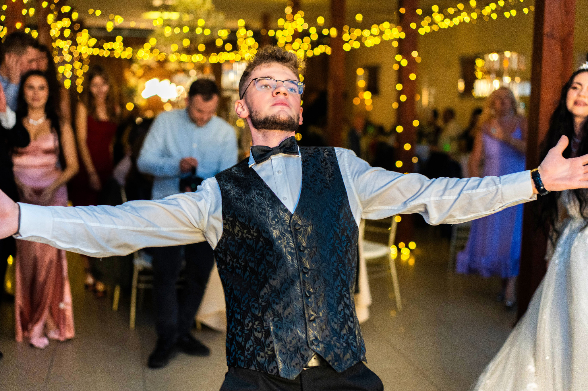 Mężczyzna w czarnej kamizelce i muszce, z rozłożonymi ramionami, na tle sali weselnej z dekoracjami świetlnymi i gośćmi.