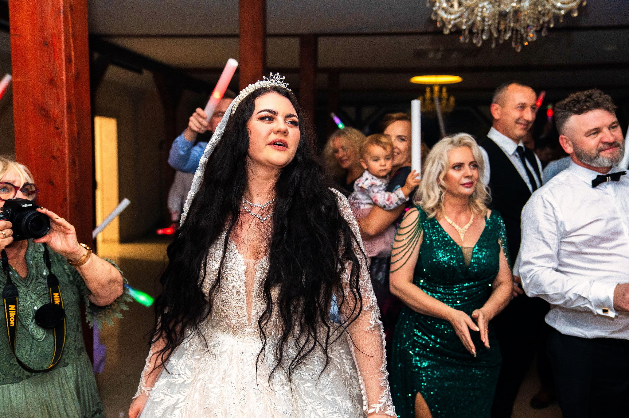 Panna młoda w koronie i bogato zdobionej sukni pozuje na weselu, otoczona gośćmi z kolorowymi, świecącymi pałeczkami; w kadrze również fotograf z aparatem Nikon.