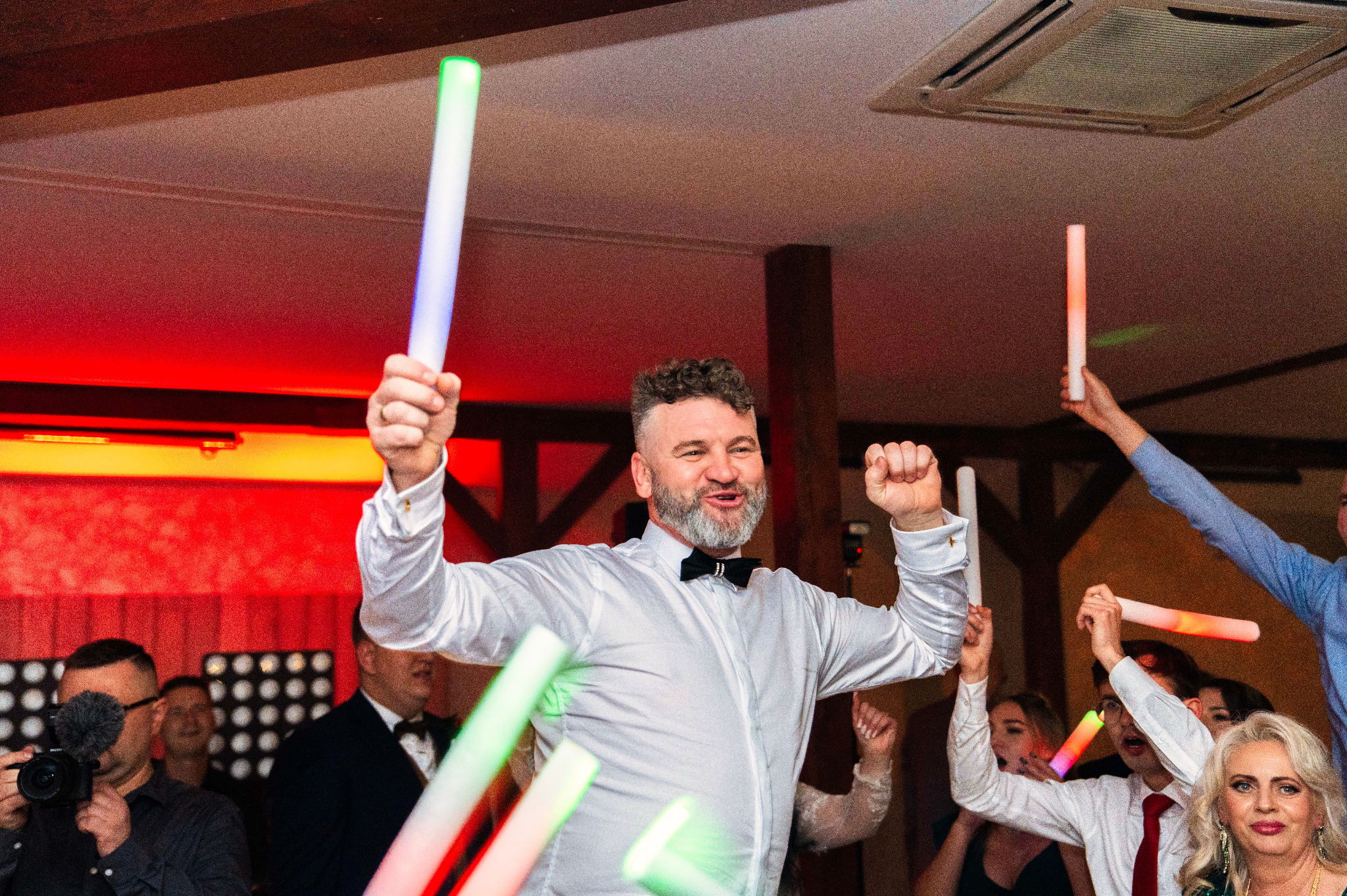 Uczestnicy wesela z podniesionymi świetlnymi pałeczkami, mężczyzna w białej koszuli i muszce w centrum kadru, w tle osoba filmująca kamerą.
