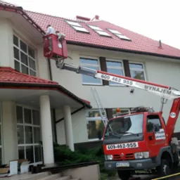 Dwóch pracowników w czerwonym koszu podnośnika teleskopowego, na tle budynku z czerwoną dachówką, wykonuje prace na wysokości, widoczny numer telefonu na ramieniu podnośnika.