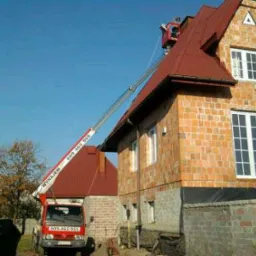 Czerwony podnośnik koszowy ustawiony przy ceglanym domu z czerwonym dachem podczas prac na wysokości, widoczne rynny i drabinki przeciwśniegowe.