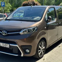 Brązowy bus Toyota Proace Verso zaparkowany na chodniku, widoczne tablice rejestracyjne z Warszawy i znak parkingu w tle.