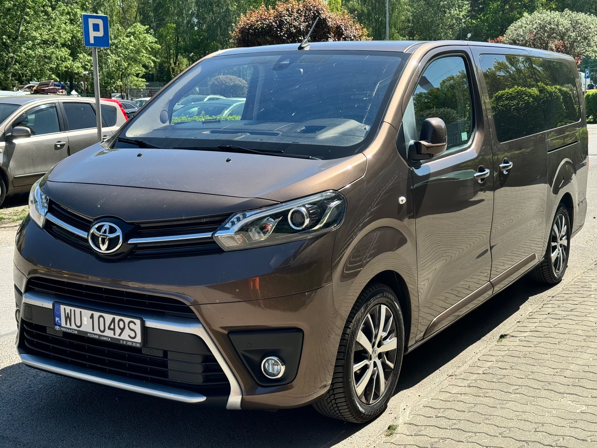 Brązowy bus Toyota Proace Verso zaparkowany na chodniku, widoczne tablice rejestracyjne z Warszawy i znak parkingu w tle.