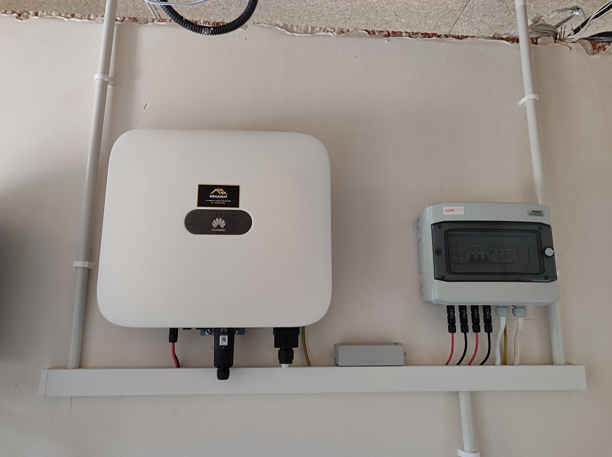 Biały inwerter Huawei z czarnym logo Megawat, zamontowany na ścianie z szarą skrzynką rozdzielczą i przewodami, widoczne elementy instalacji fotowoltaicznej.