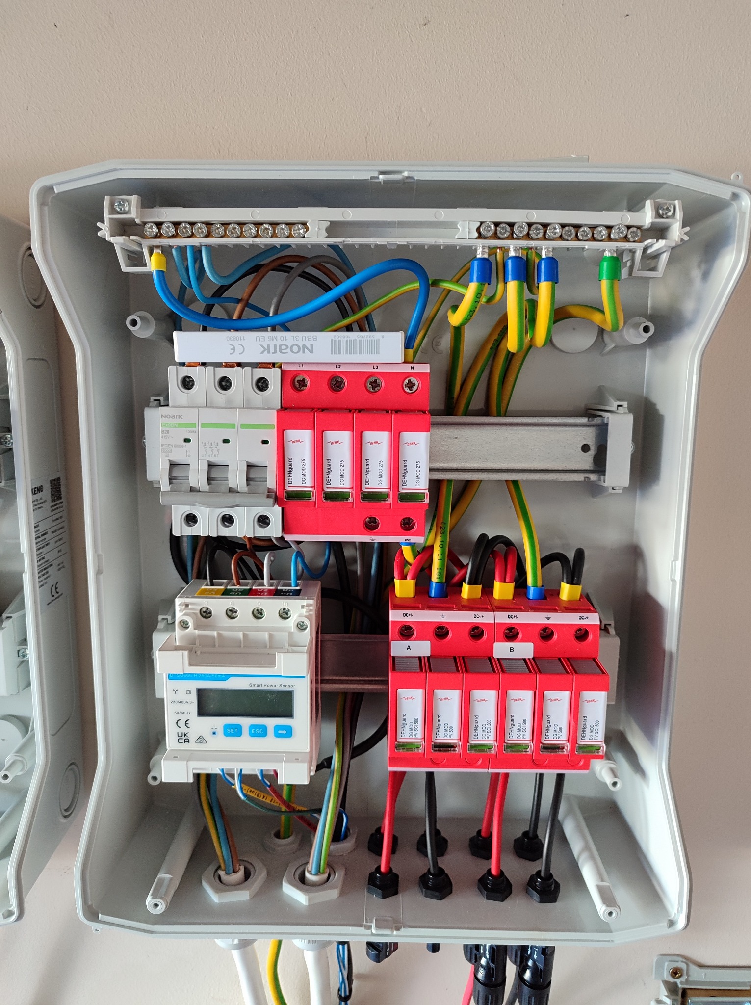 Otwarta skrzynka rozdzielcza z zainstalowanymi zabezpieczeniami przeciwprzepięciowymi DEHNguard, wyłącznikiem różnicowoprądowym Noark Ex9BN B20 i licznikiem energii Smart Power Sensor, widoczne...