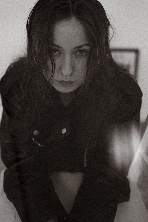 Portret kobiety w czarnej skórzanej kurtce, z mokrymi włosami, w monochromatycznej tonacji, z delikatnym efektem rozmycia w dolnej części kadru.