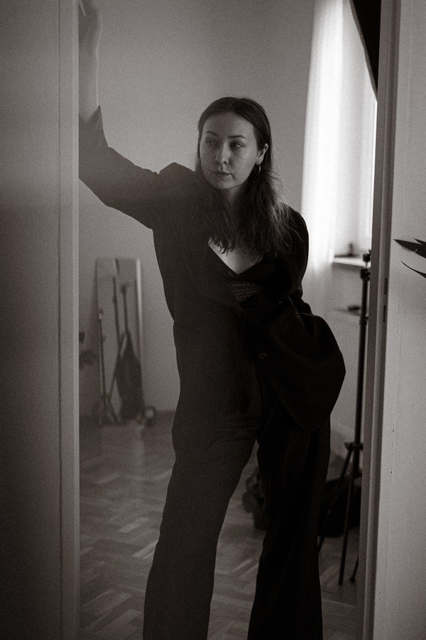 Portret kobiety w czarnym garniturze, opierającej się o framugę drzwi, z widocznym oświetleniem studyjnym w tle, utrzymany w monochromatycznej stylistyce.