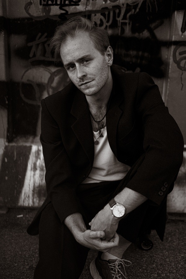 Portret mężczyzny w marynarce, siedzącego na tle ściany z graffiti, z zegarkiem na nadgarstku i naszyjnikiem na szyi, w czerni i bieli.