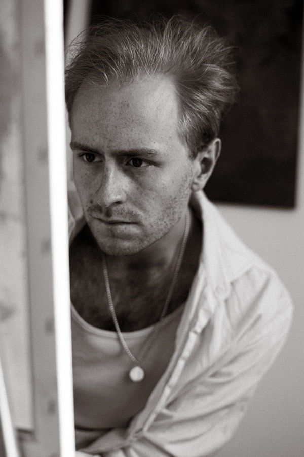 Czarno-białe portretowe ujęcie młodego mężczyzny z piegami, w rozpiętej koszuli, z medalionem na szyi, spoglądającego w lewo, z fragmentem jasnej ramy po lewej stronie.