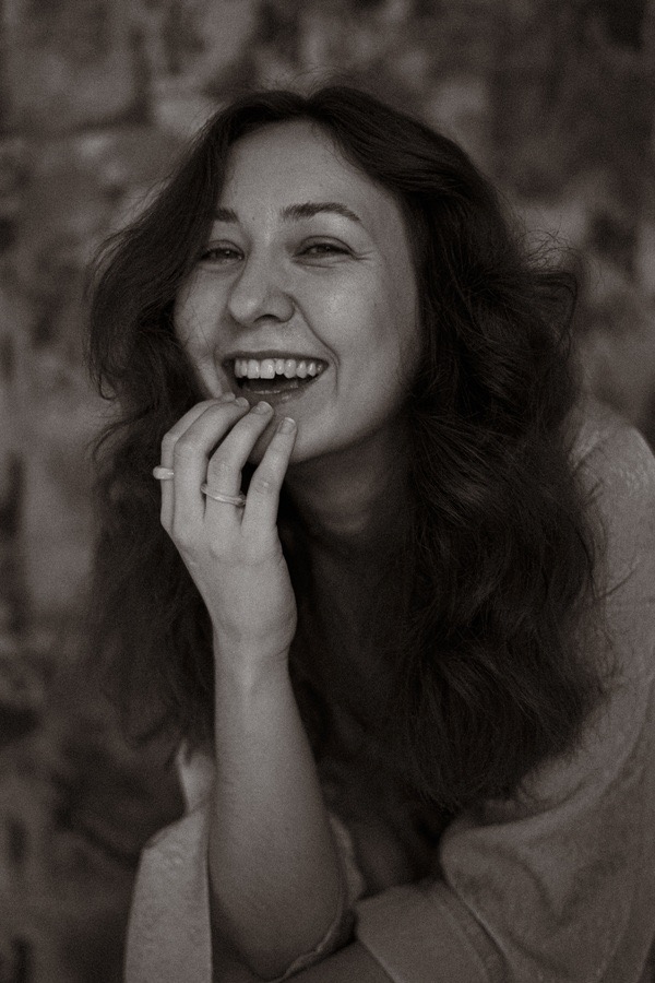 Portret uśmiechniętej kobiety w sepii, z ręką przy twarzy i dwoma pierścionkami na palcach.