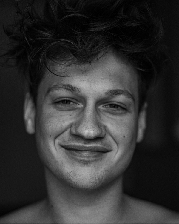 Portret młodego mężczyzny z rozwichrzonymi włosami, uśmiechającego się lekko, w czerni i bieli.