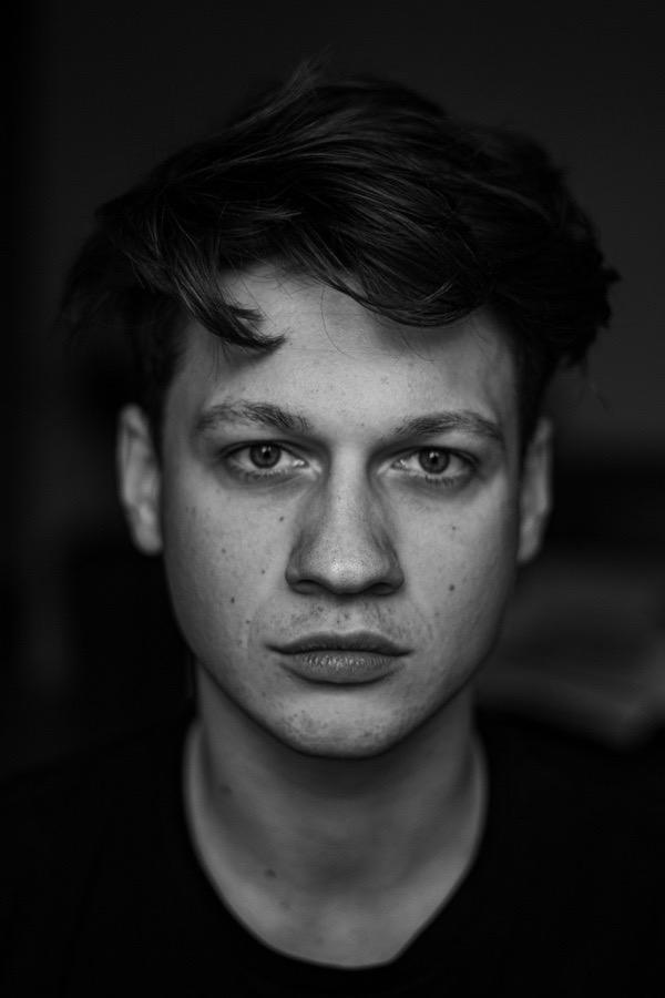 Czarno-biały portret młodego mężczyzny z krótkimi włosami i poważnym wyrazem twarzy, oświetlonego z góry, z widocznymi piegami na nosie i policzkach.