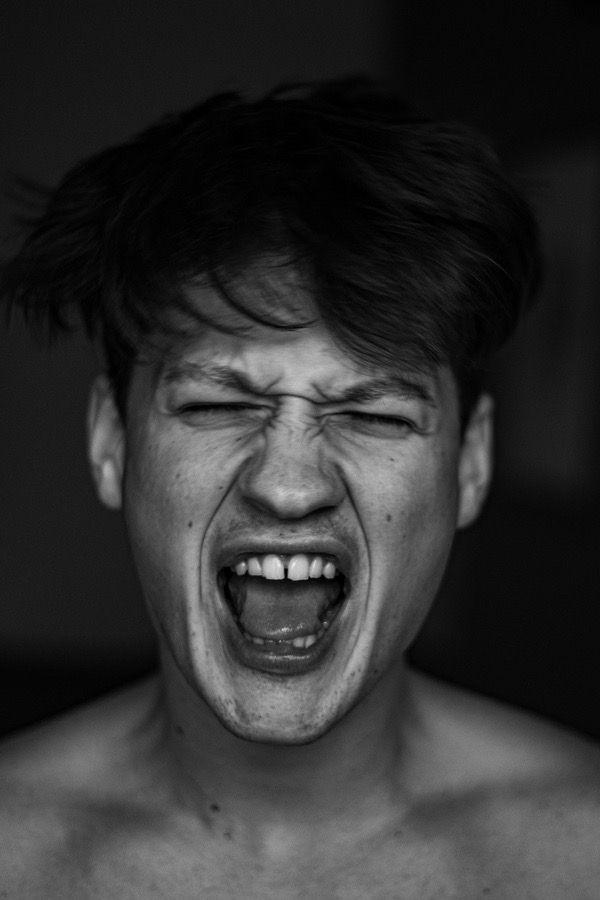 Czarno-białe zbliżenie twarzy krzyczącego mężczyzny z zamkniętymi oczami, zmarszczonym czołem i otwartymi ustami, ukazujące język i nierówne zęby.