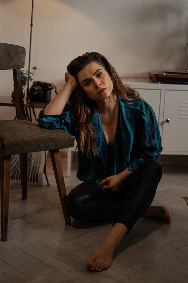 Portret kobiety siedzącej na podłodze w koszuli w zielono-niebieskie paski, opierającej głowę na krześle, z gramofonem i szafką w tle.