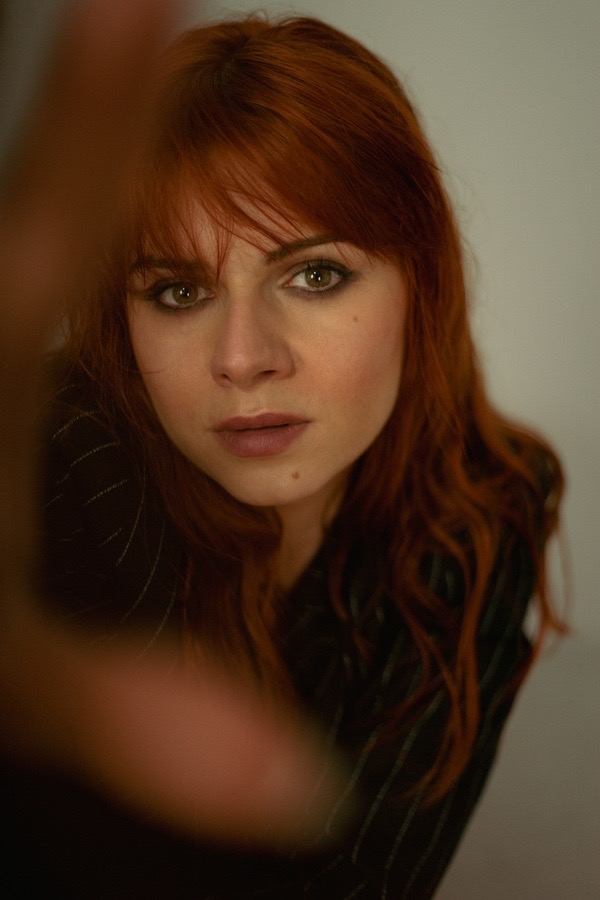 Portret kobiety z rudymi włosami, zielonymi oczami i delikatnym makijażem, ujęcie z dołu z częściowo zasłoniętą twarzą przez rozmazaną dłoń w pierwszym planie.