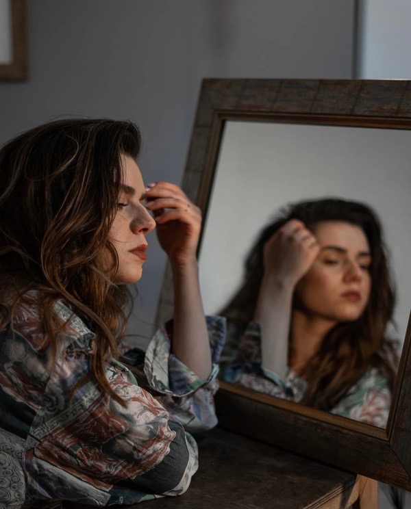 Kobieta w koszuli z wzorem spogląda w lustro z drewnianą ramą, dotykając włosów.