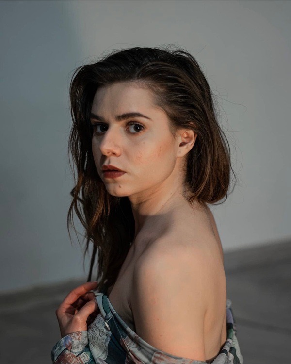 Portret kobiety z mokrymi włosami, spoglądającej w bok, z delikatnym makijażem i okrytej wzorzystą tkaniną na tle szarej ściany.