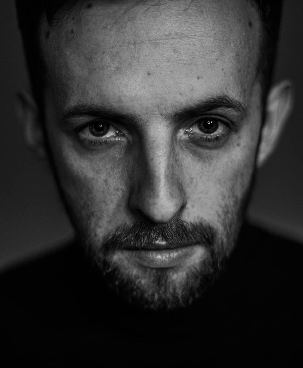 Czarno-biały portret mężczyzny z lekkim zarostem, wpatrującego się w obiektyw, z ciemnym tłem.