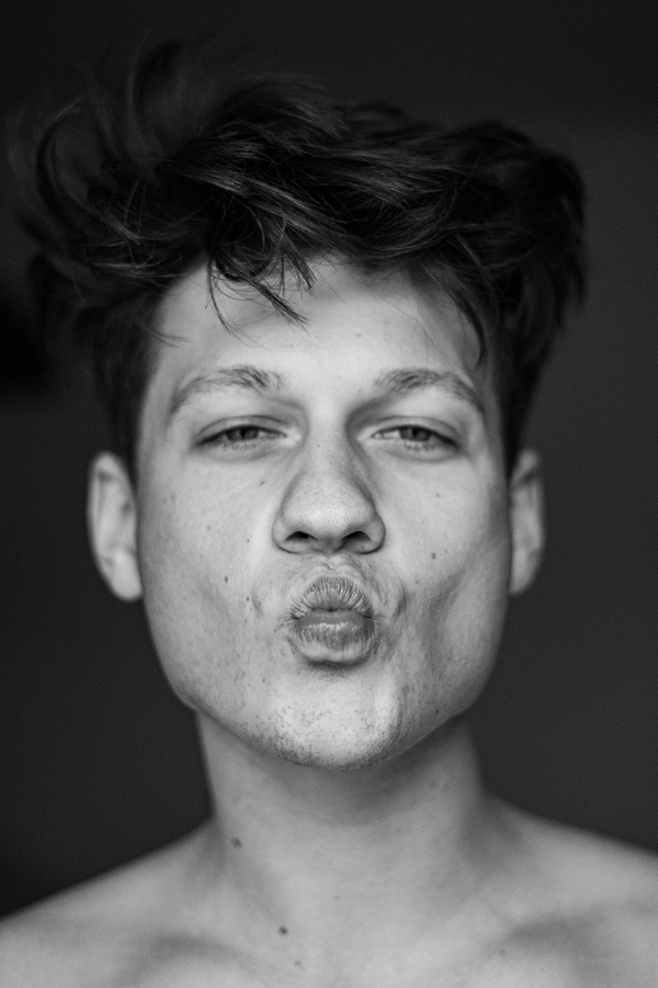 Czarno-biały portret młodego mężczyzny z zmierzwionymi włosami, robiącego dzióbek ustami.